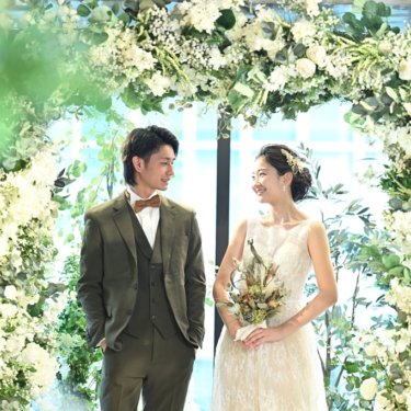 Couple standing beneath a white floral archway, groom in an olive three-piece suit and bride in a lace gown holding a wildflower bouquet.