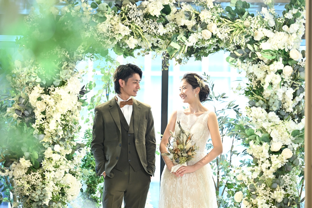Couple standing beneath a white floral archway, groom in an olive three-piece suit and bride in a lace gown holding a wildflower bouquet.