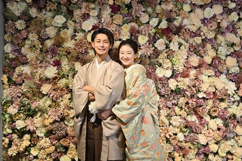 Couple smiling in front of a dense antique-toned flower wall, groom in a beige kimono and bride in pale green floral kimono.
