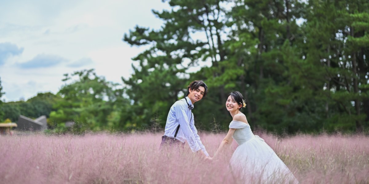 Couple walking hand in hand through a pink field of blooming pampas-like grass at Nagai Park, glancing back with a smile.