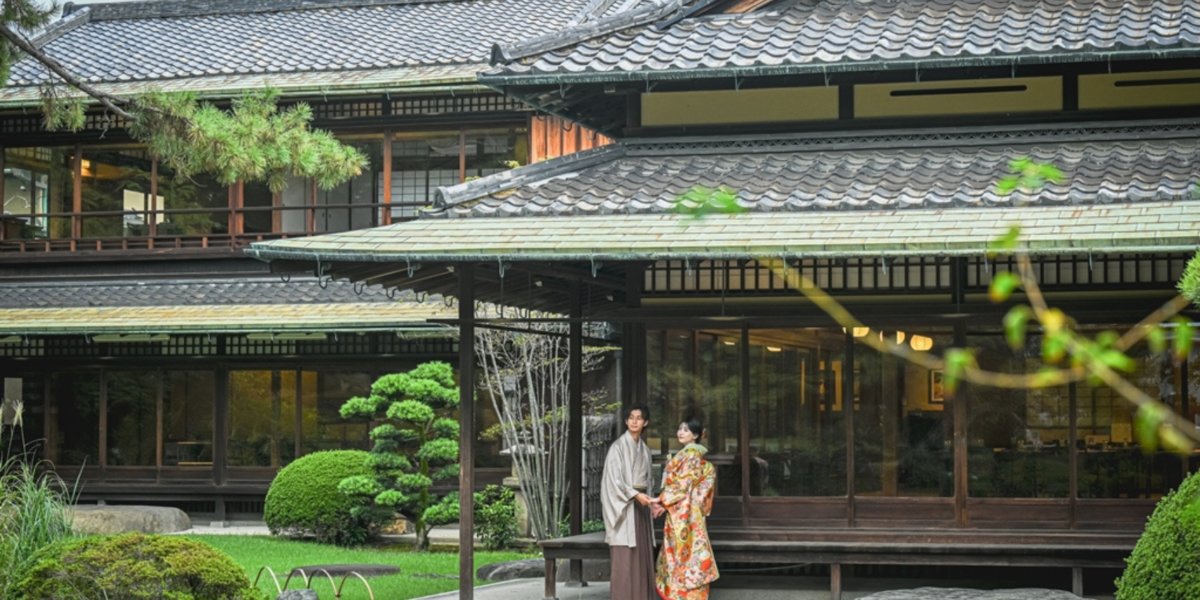 Couple in kimono standing before the grand traditional estate of Gofuso, its sweeping tiled roofs and manicured pines behind them.