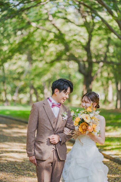 Groom in a brown tweed suit with a pink bow tie and bride in a ruffled OB20 wedding dress laughing arm in arm during a park wedding photoshoot