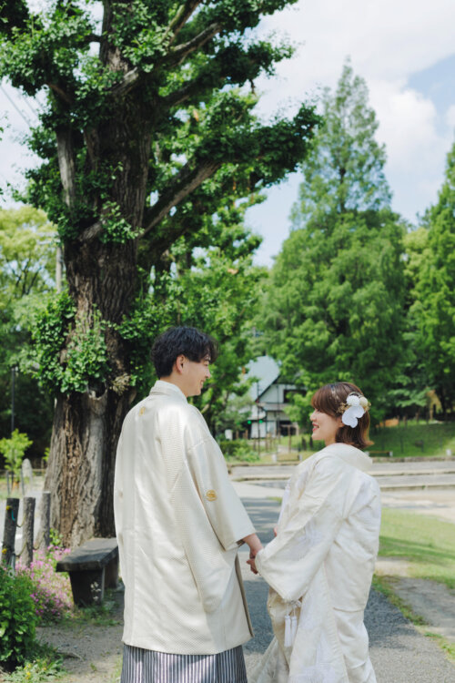 Groom in white montsuki hakama and bride in shiromuku holding hands under a large tree in a green park, a pre-wedding photoshoot in Japan