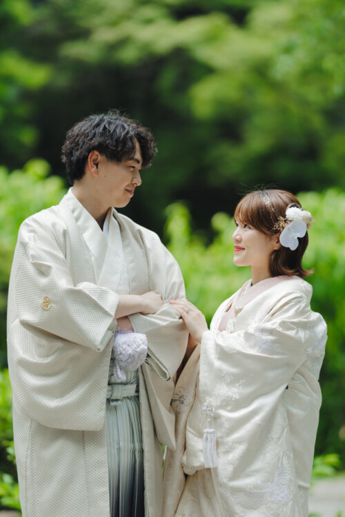 Groom in white montsuki hakama and bride in shiromuku with a pink collar gazing at each other against fresh greenery, kimono wedding photos in Japan