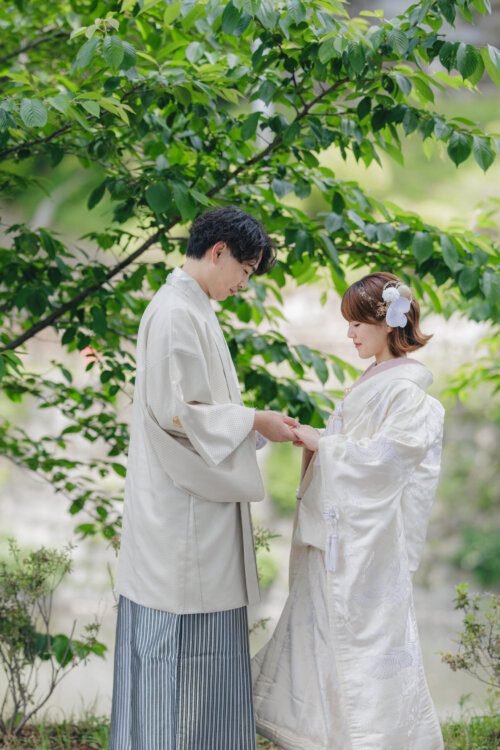 Groom in white montsuki hakama and bride in shiromuku holding hands under leafy branches in early summer, Japan wedding photography