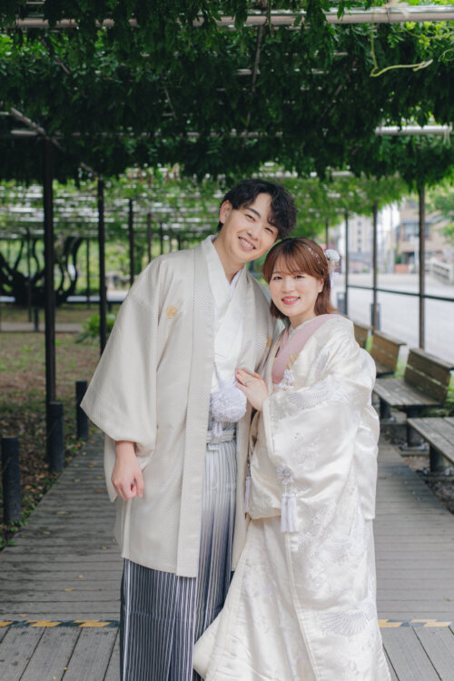 Smiling groom in white montsuki hakama and bride in shiromuku snuggling on a boardwalk under a wisteria trellis, kimono wedding photos in Japan