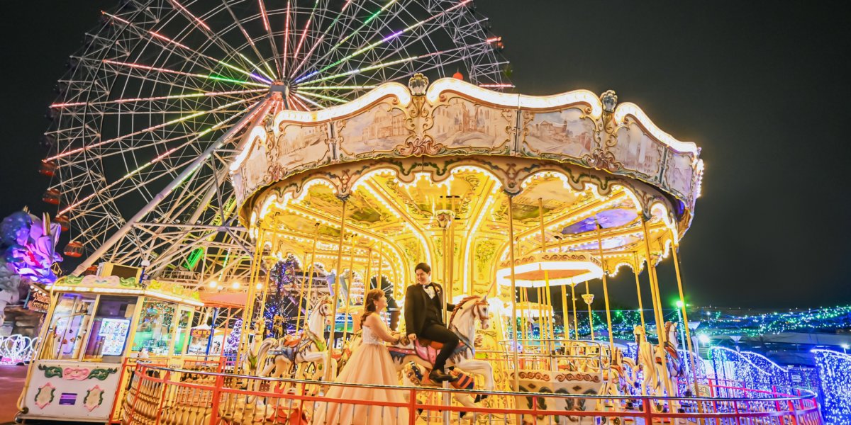 Couple riding a carousel horse at night, a giant illuminated Ferris wheel behind them at Sea Train Land.