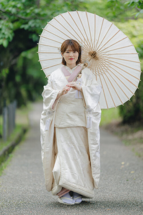 Bride in the SF105 shiromuku with a pink underkimono holding a wagasa umbrella on a shaded path, a full-length pre-wedding photoshoot in Japan