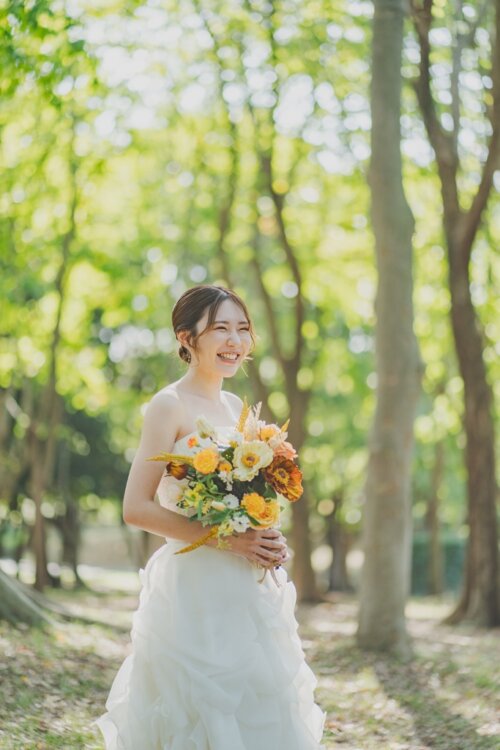 Bride in a strapless ruffled wedding dress smiling with an orange and yellow bouquet among tall trees, a pre-wedding portrait in Japan