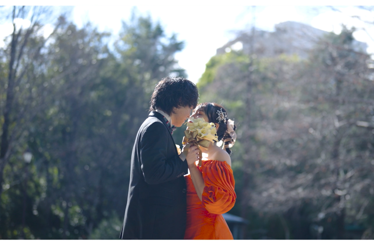 Video still of the couple sharing a kiss behind a bouquet of white calla lilies, sunlit trees behind them.