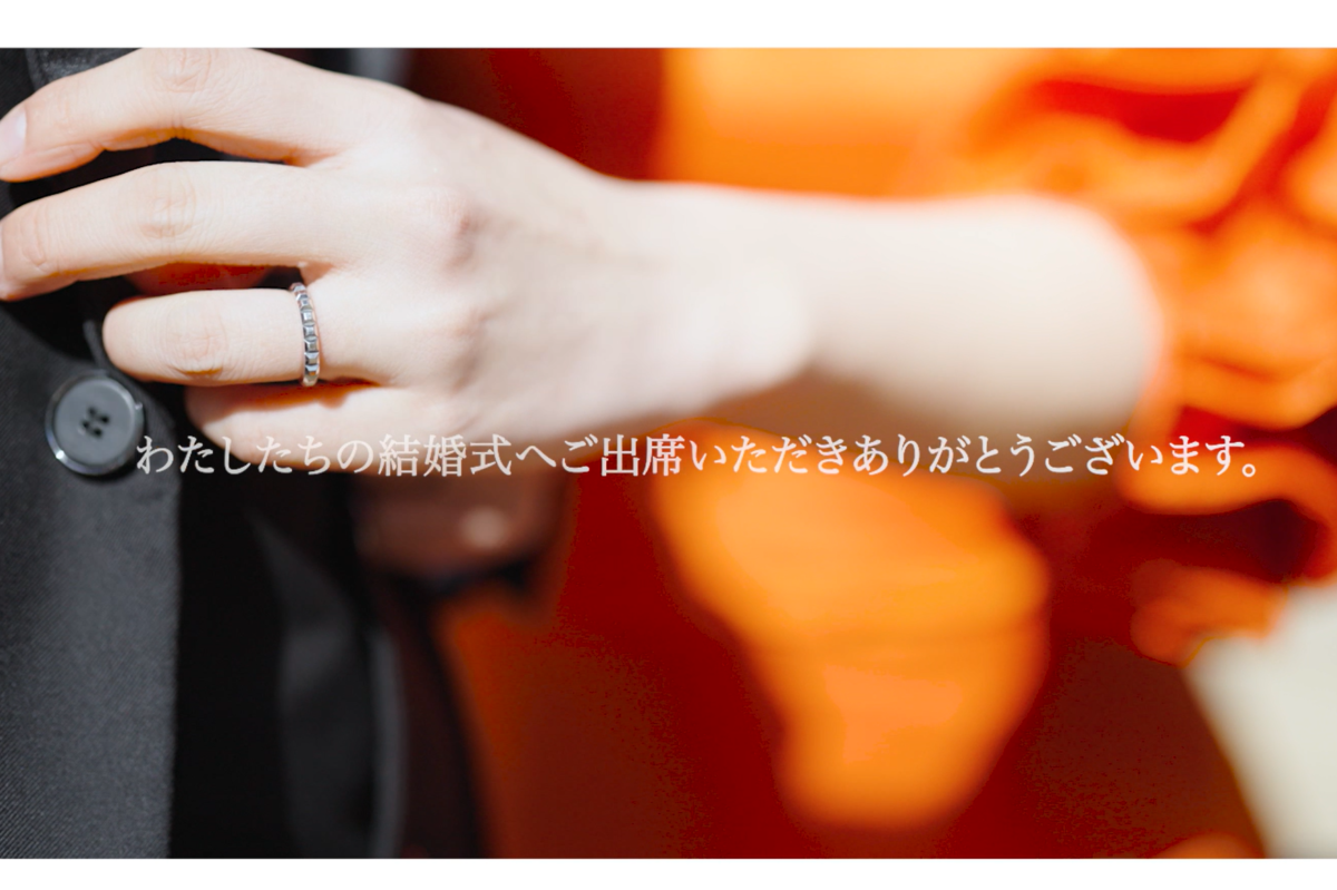 Close-up video still of a hand with a wedding ring resting on the groom's black suit, with Japanese text thanking guests for attending the wedding.