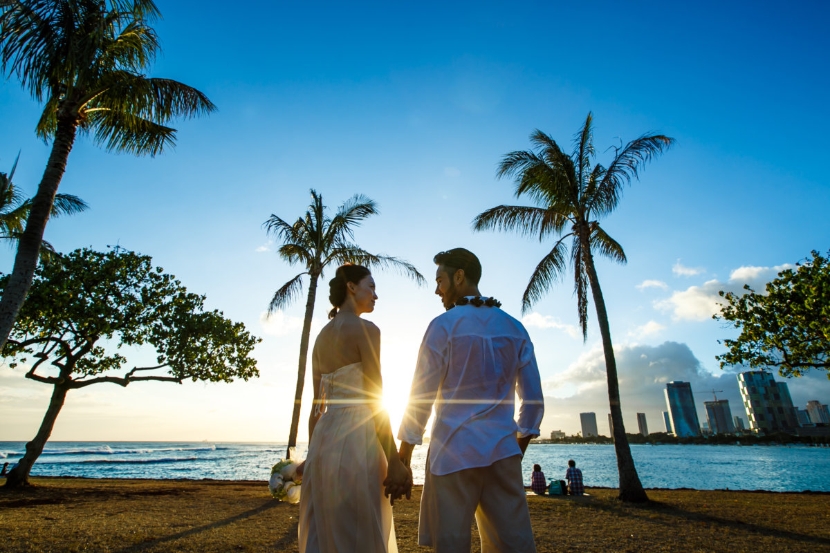 Couple holding hands on a Hawaii beach at sunset between palm trees with the city skyline beyond, a honeymoon photo wedding