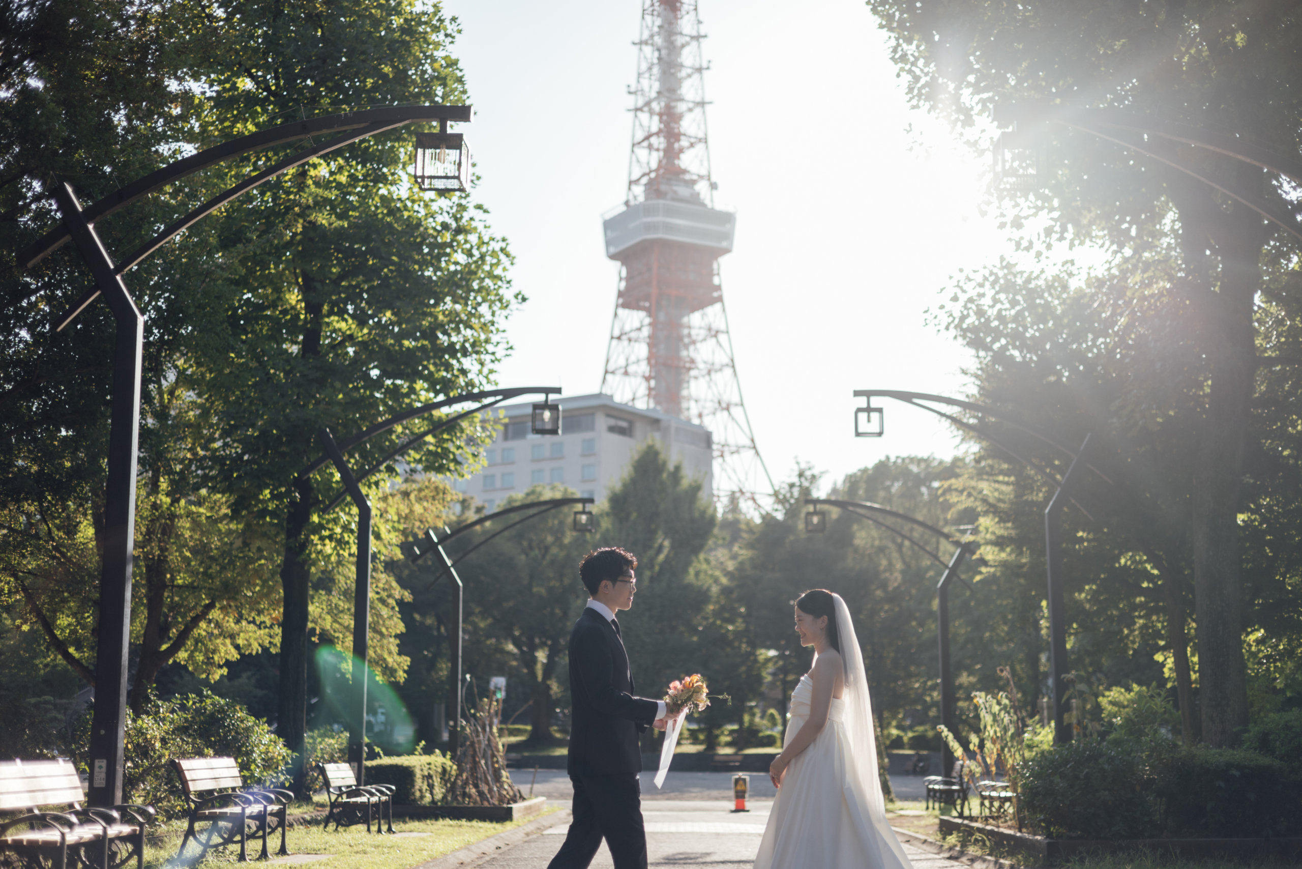 Approaching each other along a sunlit park path, a famous red-and-white broadcast tower rising in the background.