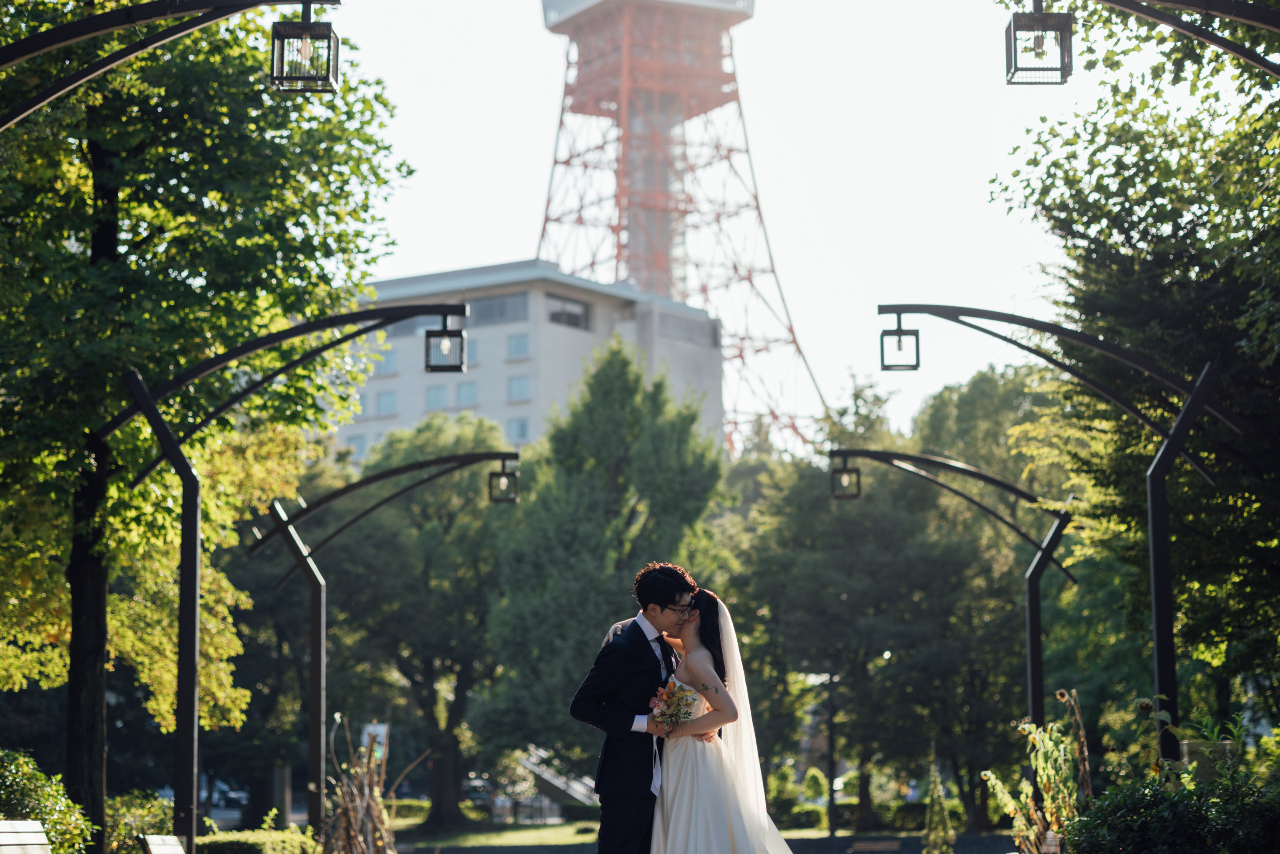 A tender kiss framed by garden archways, Tokyo Tower standing tall behind the trees.