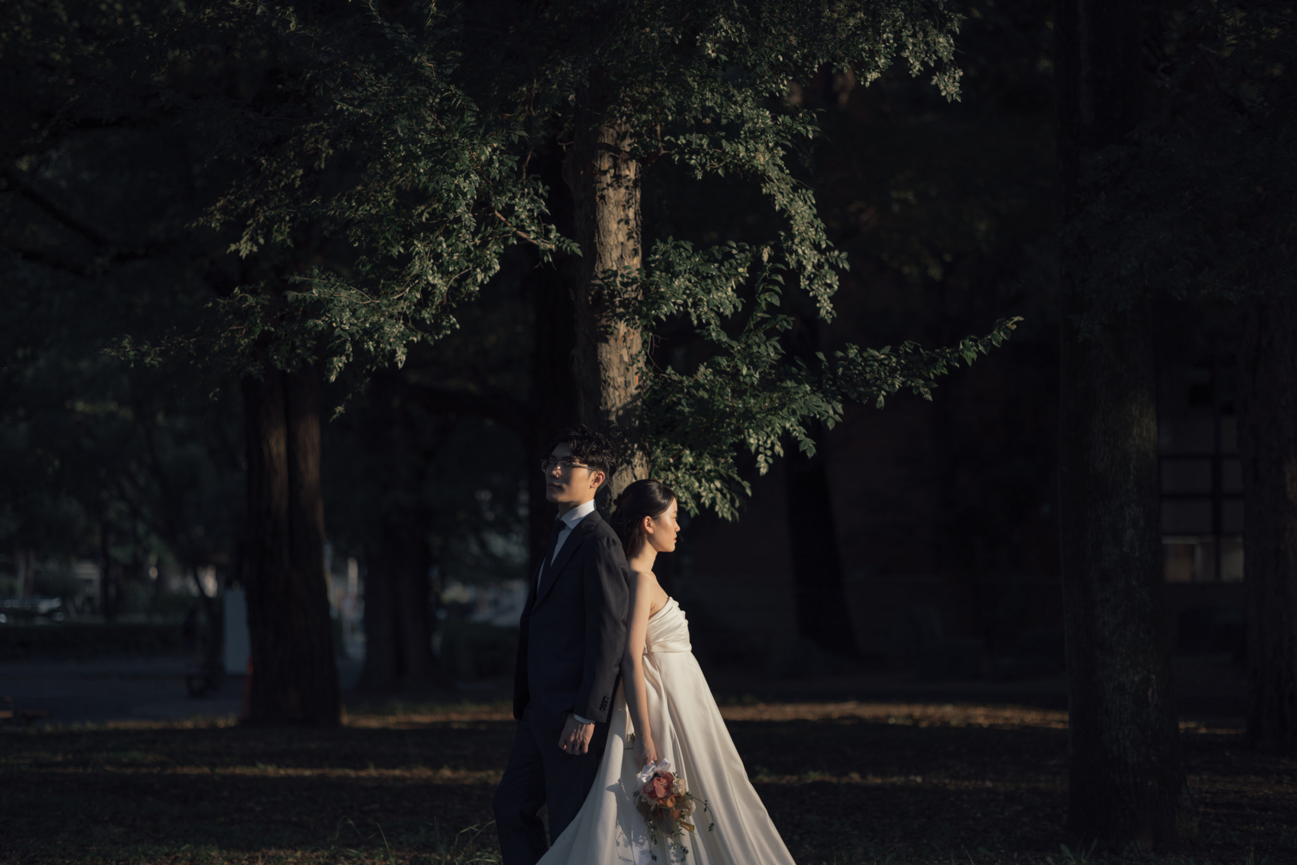Standing back to back in dappled shade beneath a towering tree, a quiet, contemplative portrait.