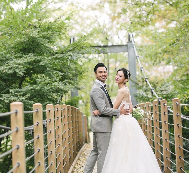 Crossing a suspension bridge through lush greenery at Kinuta Park, the groom glances back with a warm smile.