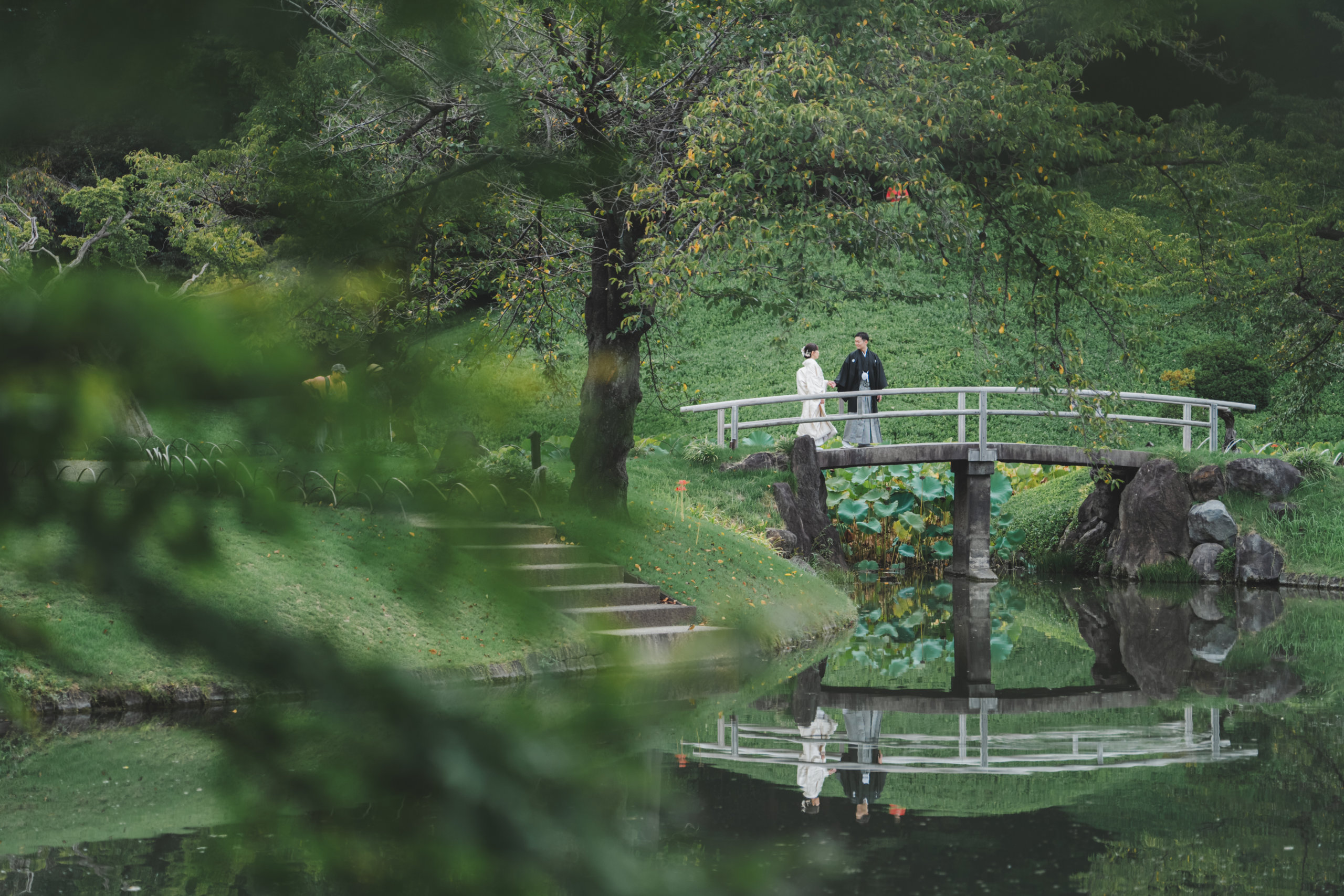 Framed by soft green foliage, the couple crosses a stone bridge over a lotus pond in the distance.