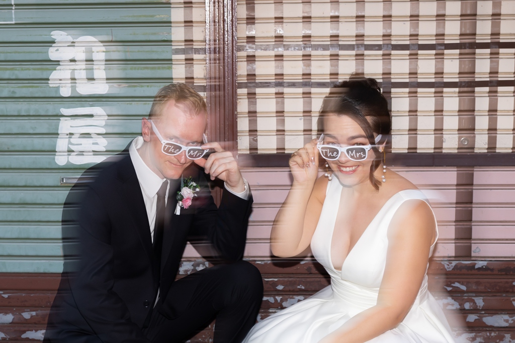 Striking a playful pose in matching 'The Mrs.' sunglasses against a vintage shop shutter.