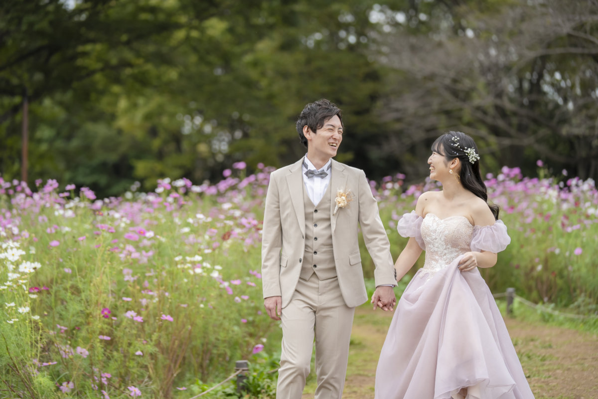 Groom in a beige three-piece suit and bride in a lavender puff-sleeve gown walking hand in hand through a cosmos field in autumn, Japan wedding photography