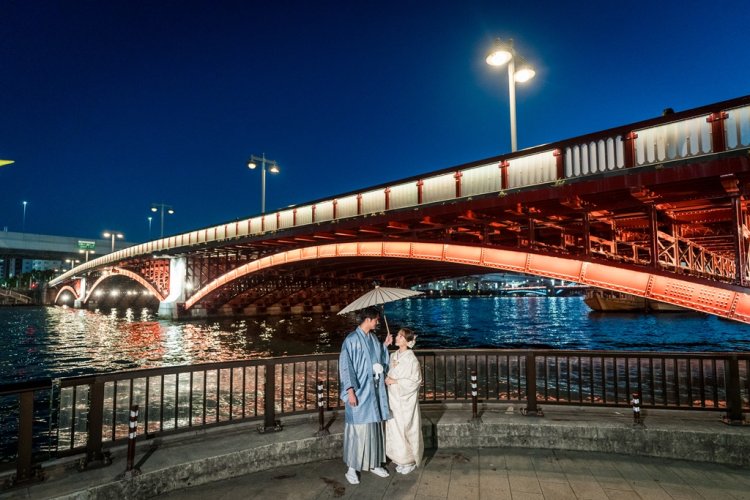 Sheltering under a single umbrella beside the glowing red Azuma Bridge over the Sumida River at night.