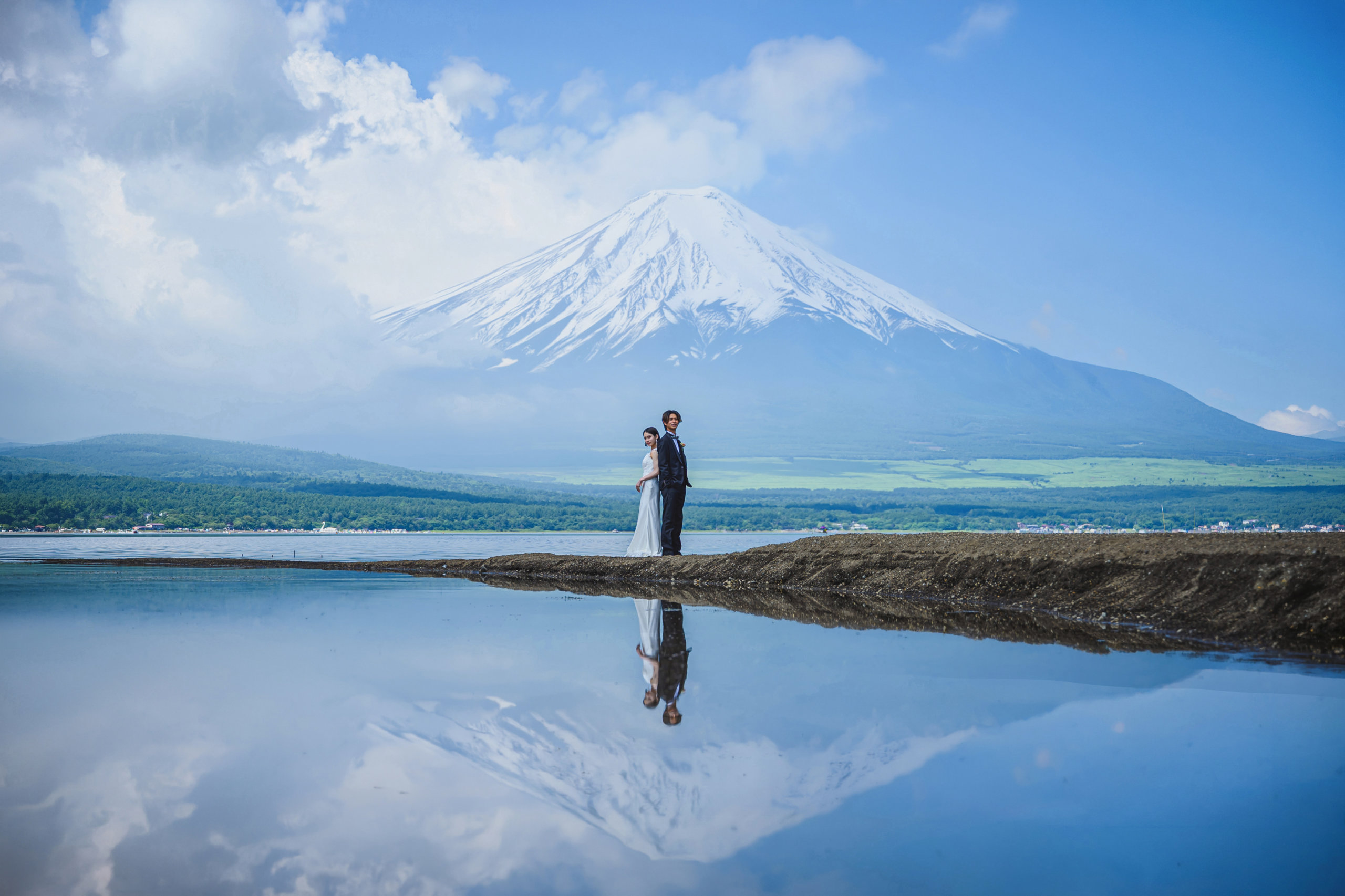 Bride and groom standing on a narrow spit of land, snow-capped Mt. Fuji perfectly mirrored in the still lake water below, a signature shot for pre-wedding photography in Japan.