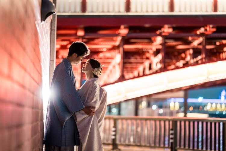 An intimate embrace beneath the illuminated arches of Azuma Bridge, warm light spilling across the Sumida riverside.