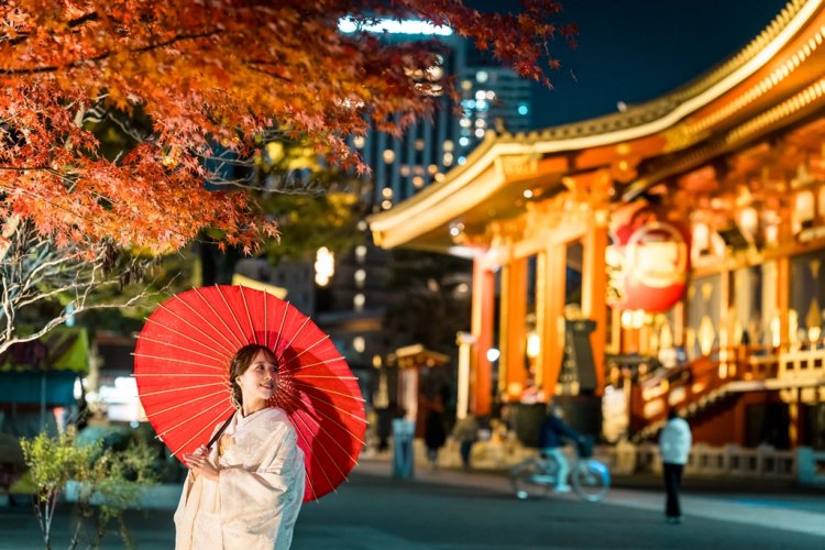 A bright red paper umbrella frames the bride beneath autumn leaves, Senso-ji Temple's glowing hall lighting up the night behind her.