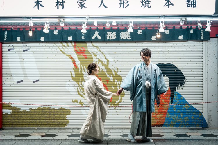 Holding hands outside a shuttered Asakusa storefront, its colorful hand-painted mural glowing under string lights.