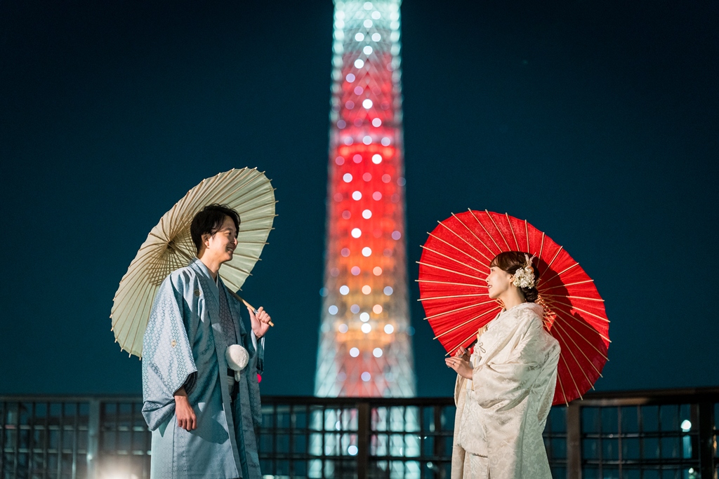 Sheltering under cream and red paper umbrellas, the illuminated Tokyo Skytree glowing brightly against the night sky.