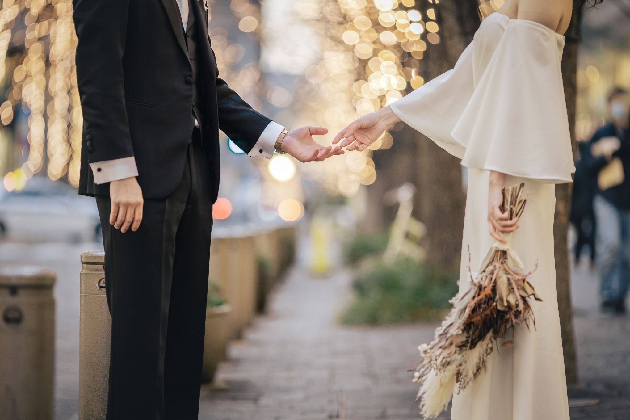 Close-up of the couple's hands reaching for each other, groom in a black tuxedo and bride in a flowing cream dress holding a dried-flower bouquet, on a tree-lined street glowing with warm bokeh lights.
