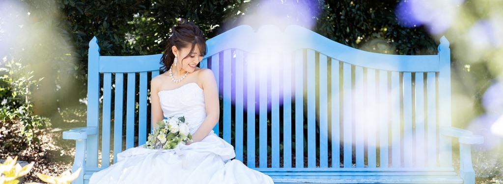 The bride sits quietly on a pale blue garden bench at Blue Bonnet, a bouquet of white roses resting in her lap.