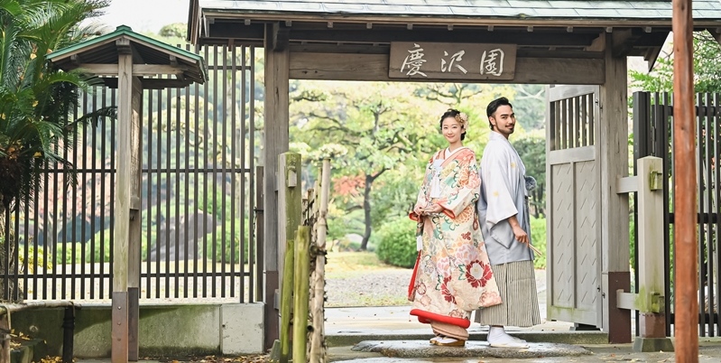 Couple in kimono standing back to back beneath a wooden gate reading 'Keitakuen', leading into a Japanese garden.