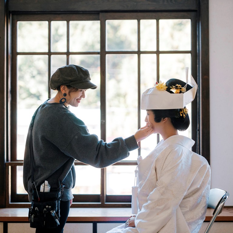 A hairstylist adjusts the bride's floral hair ornaments beneath her white tsunokakushi headpiece.