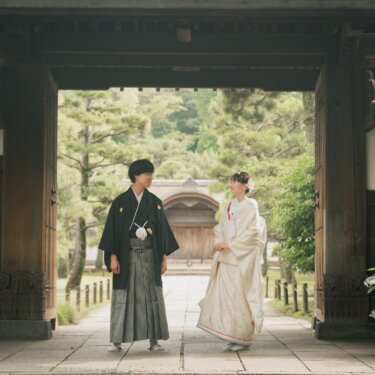 Couple in shiromuku and montsuki standing beneath a large wooden temple gate with hydrangeas in bloom, a serene Japan wedding photography scene