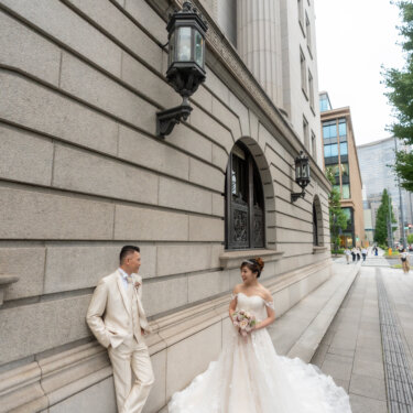 Groom leaning against the granite wall as his bride stands facing him with her train spread over the sidewalk, a relaxed pose for a pre-wedding photoshoot in Tokyo.