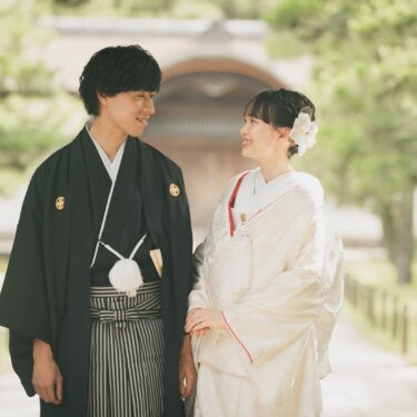 Bride and groom in traditional wedding kimono smiling at each other on a temple path lined with pine trees seen in this kimono wedding photos in Japan