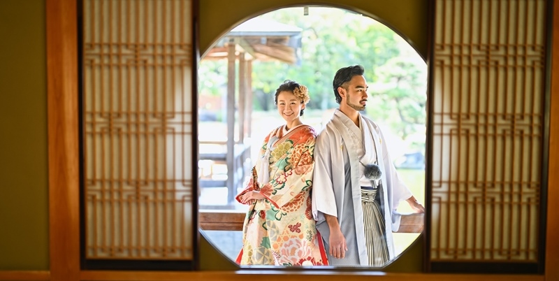 Couple in kimono framed inside a round window, garden and traditional building visible through the opening at Daisen Park.