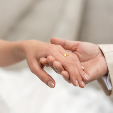 Close-up of the groom gently holding his bride's hand to show her gold wedding ring against the soft white of her gown, a delicate detail shot for wedding photos.