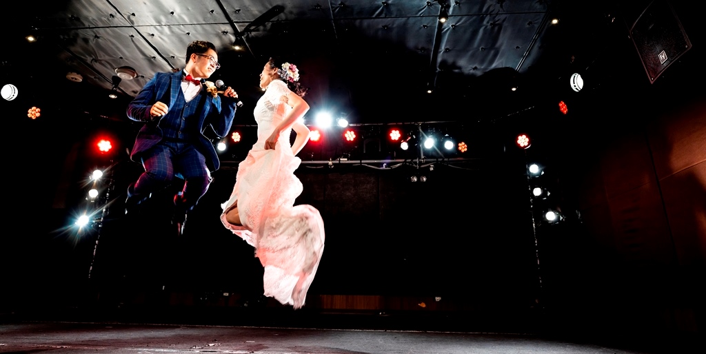 Leaping mid-air on a dazzling concert stage, microphone in hand, surrounded by vibrant stage lighting at a Nagoya live music venue.