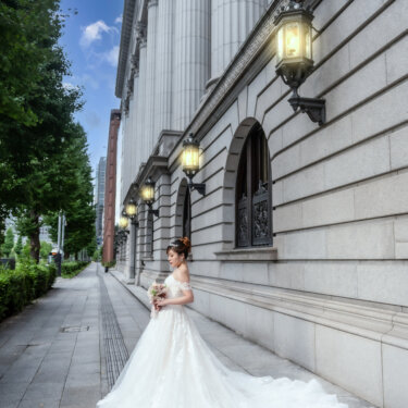 Bride standing alone beside a stone building lined with lanterns under a bright blue sky, her long lace train fanned out on the Marunouchi pavement for her pre-wedding photos.