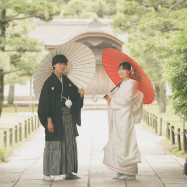 Couple in wedding kimono holding cream and red wagasa parasols in a temple gateway, the stone approach and traditional roof behind them