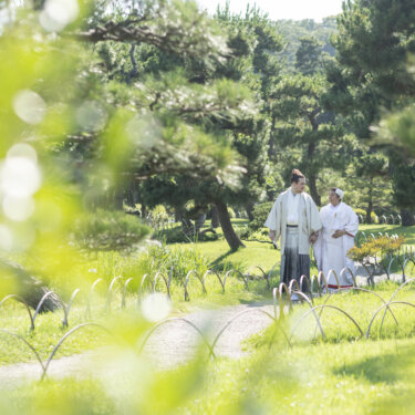 Couple in white kimono strolling along a pine-lined lawn with soft bokeh from green leaves in the foreground, a dreamy shot from their pre-wedding photoshoot in Yokohama