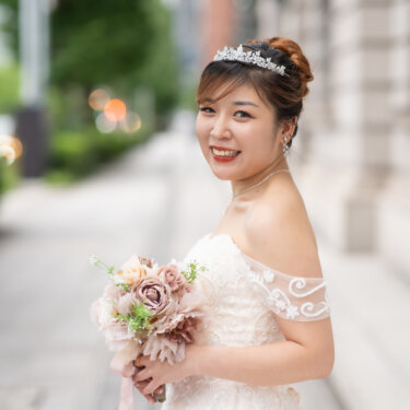 Portrait of the bride in an off-shoulder lace gown and sparkling tiara, holding a dusty-pink bouquet and glancing back on a tree-lined sidewalk, a soft moment from her wedding photos.
