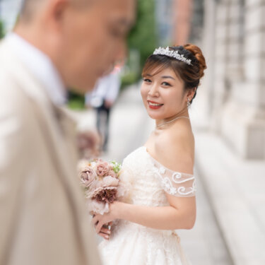 Bride looking back over her shoulder with a smile, framed by the out-of-focus groom in the foreground beside a historic stone building in Marunouchi, a creative idea for wedding photography.