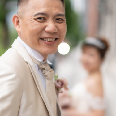 Groom in a champagne tuxedo with a pink boutonniere smiling over his shoulder, his bride softly blurred behind him on a Tokyo street during their pre-wedding photoshoot.
