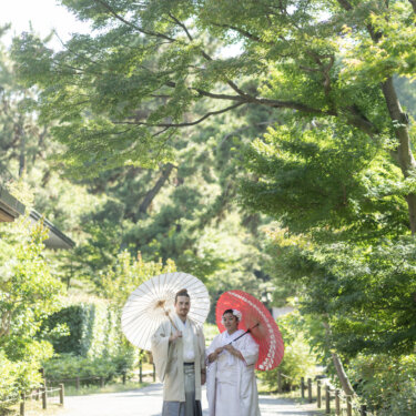 Couple in shiromuku and hakama walking under white and red wagasa umbrellas along a leafy garden path at Sankeien Garden, a classic Japan wedding photography scene