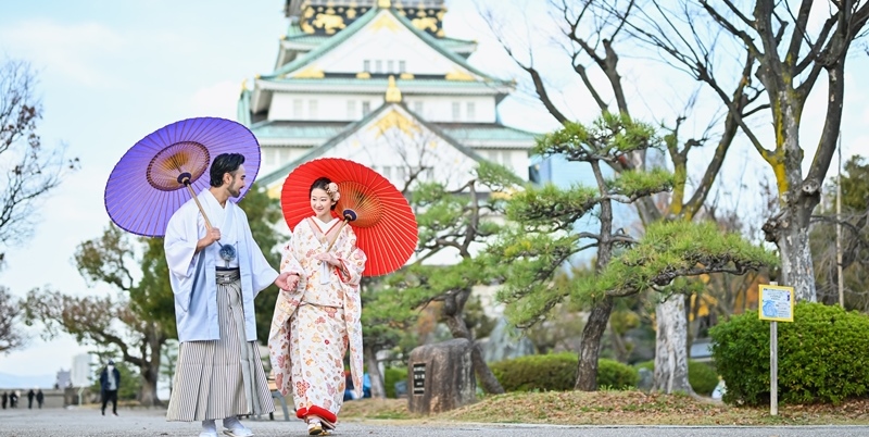Couple in kimono holding purple and red paper umbrellas walking a path with Osaka Castle's golden gables rising behind them.