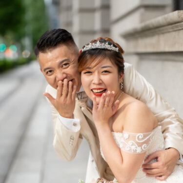 Playful couple covering their mouths with their hands and laughing toward the camera on a Marunouchi sidewalk, a fun and cheerful pose for pre-wedding photos.