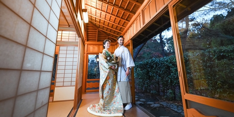 Couple in kimono standing in a wooden corridor, bride holding a bouquet, a garden visible beyond the sliding shoji doors at Murasaki-tei.