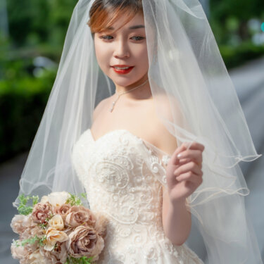 Bride in a sweetheart lace gown, tiara and sheer veil lifting the veil edge with a gentle smile, holding a mauve rose bouquet on a leafy street in this bridal wedding photo.